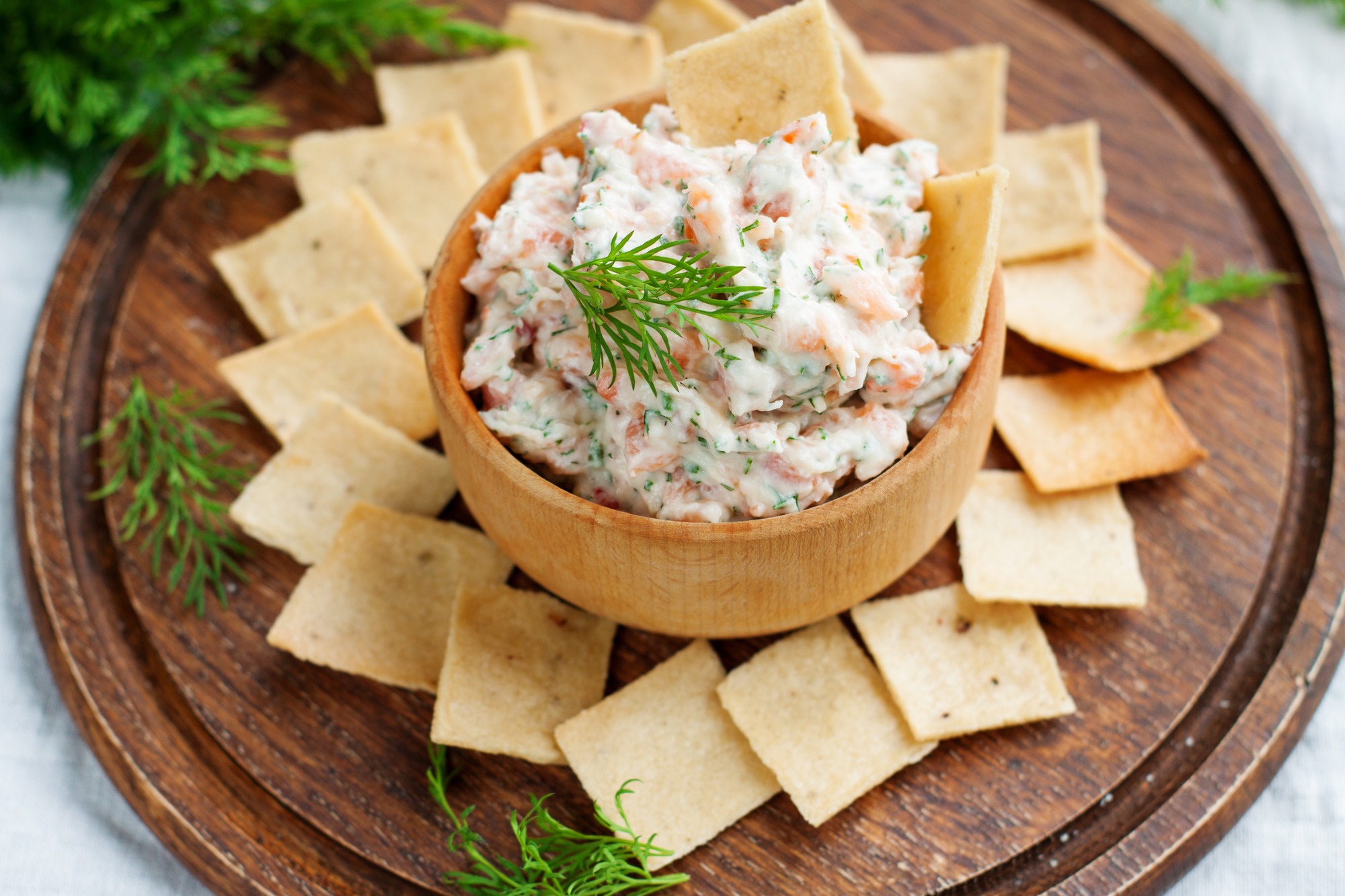 Smoked salmon, cream cheese and dill dip in wooden bowl served with gluten free crackers on round cutting board